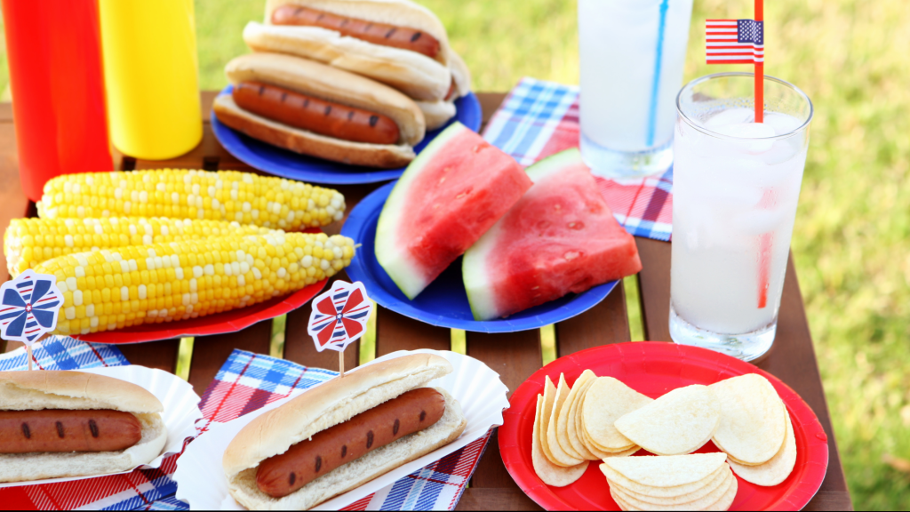 A display of common picnic foods like hot dogs, corn on the cob, watermelon, and chips on a picnic table cloth with Fourth of July accessories.